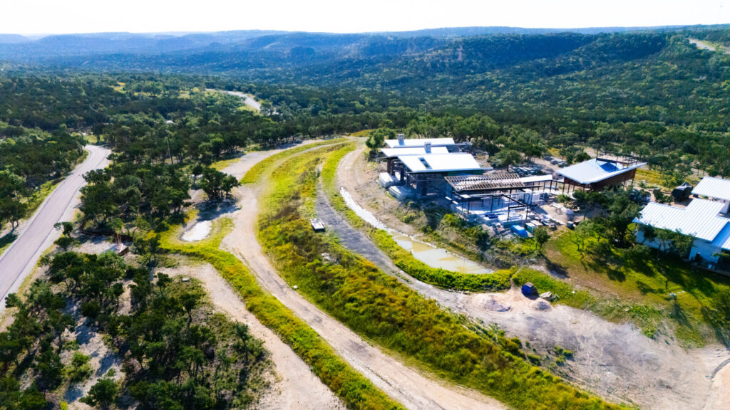 Aerial view of a Central Texas property with large contour berms, water harvesting earthworks, and native vegetation surrounding a modern homestead, showing an example of regenerative permaculture landscape design.