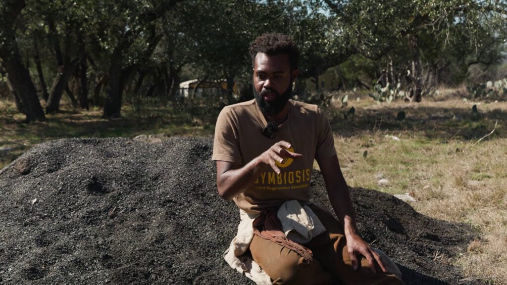 Symbiosis crew member holding a handful of biochar beside a large pile of it in the Texas Hill Country