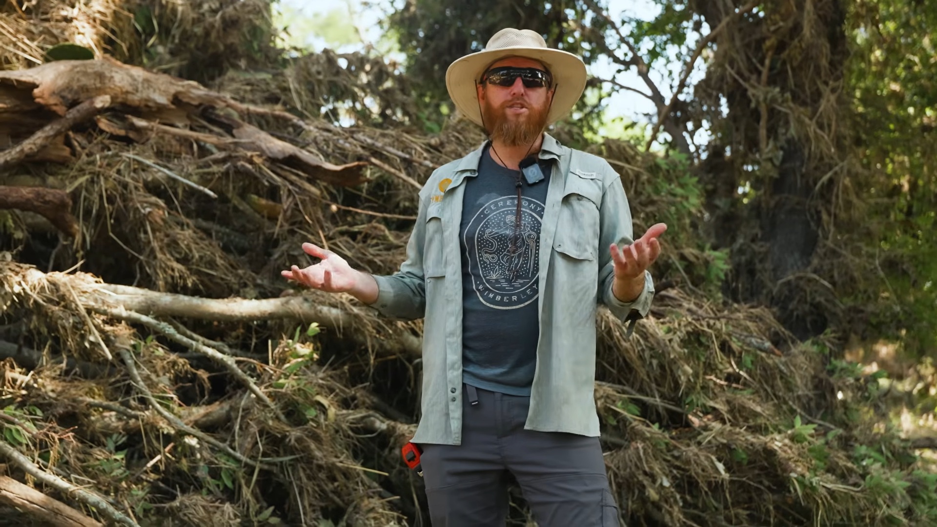 A Symbiosis team member standing beside a large root wad and pile of flood debris on Central Texas land, showing the scale of damage a landowner needs to document.
